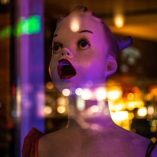 A child-like mannequin with makeup and an open mouth stares out a store window into the night. Lights reflect off the window.