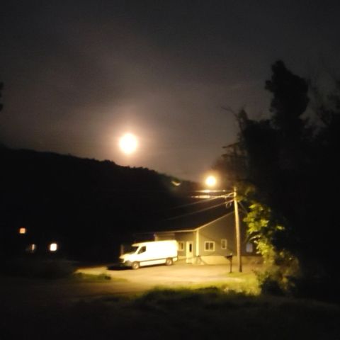 A night sky with a full moon fills half the frame. The other half is a small warehouse with a plain white van backed up to it. A street light illuminates the warehouse and the van