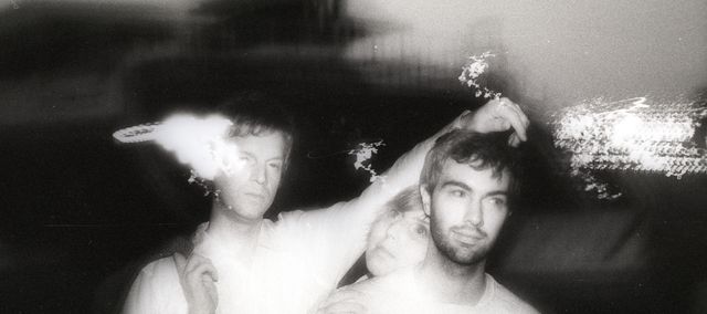 A scan of a black and white printed photo. A long exposure causes flits of light around the band. The three band members are huddled together in an active pose.