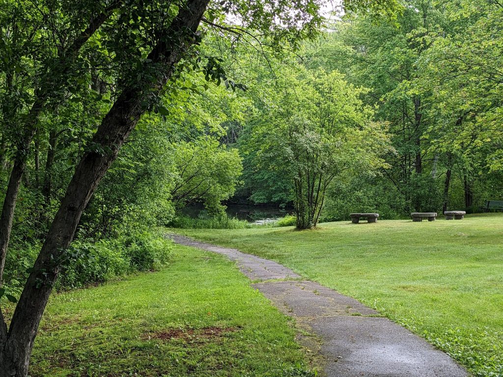 A path leads past a grassy field with granite benches toward a pond 