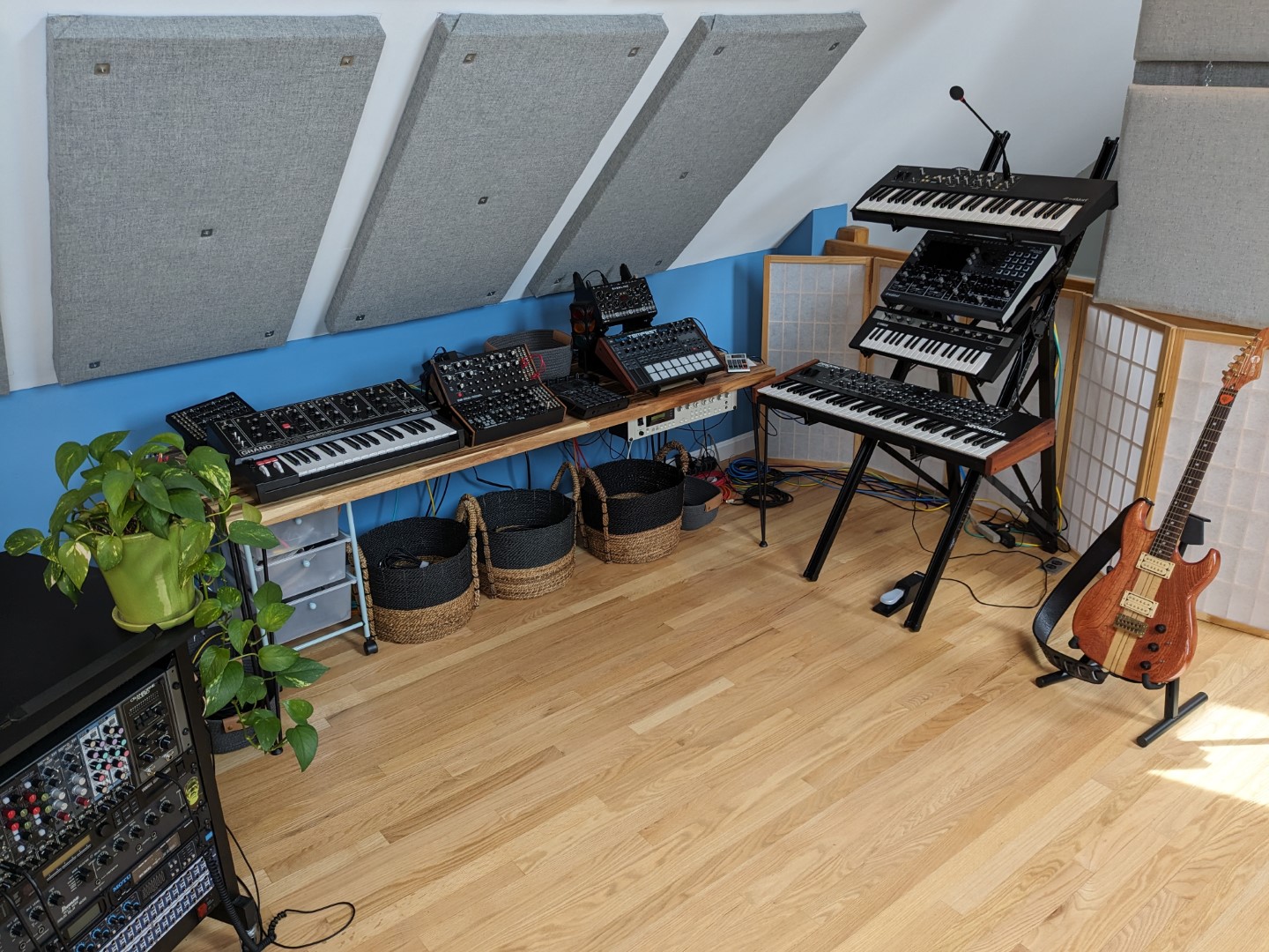 A desk and stand full of synths, a rack full of music gear, and an electric guitar