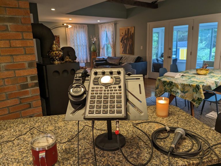 A Roland SP404 MK2 sits on a kitchen counter alongside a mic, headphones, coffee, and a lit candle.