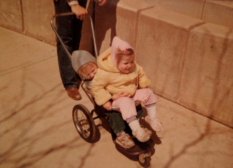 Two very small children share a single small stroller in the early 80s.