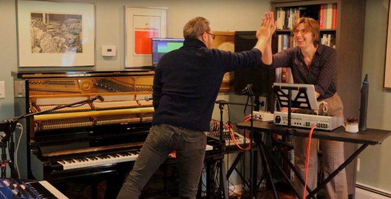 Greg and Alison high five in a living room with a piano and a bunch of other musical instruments around them.