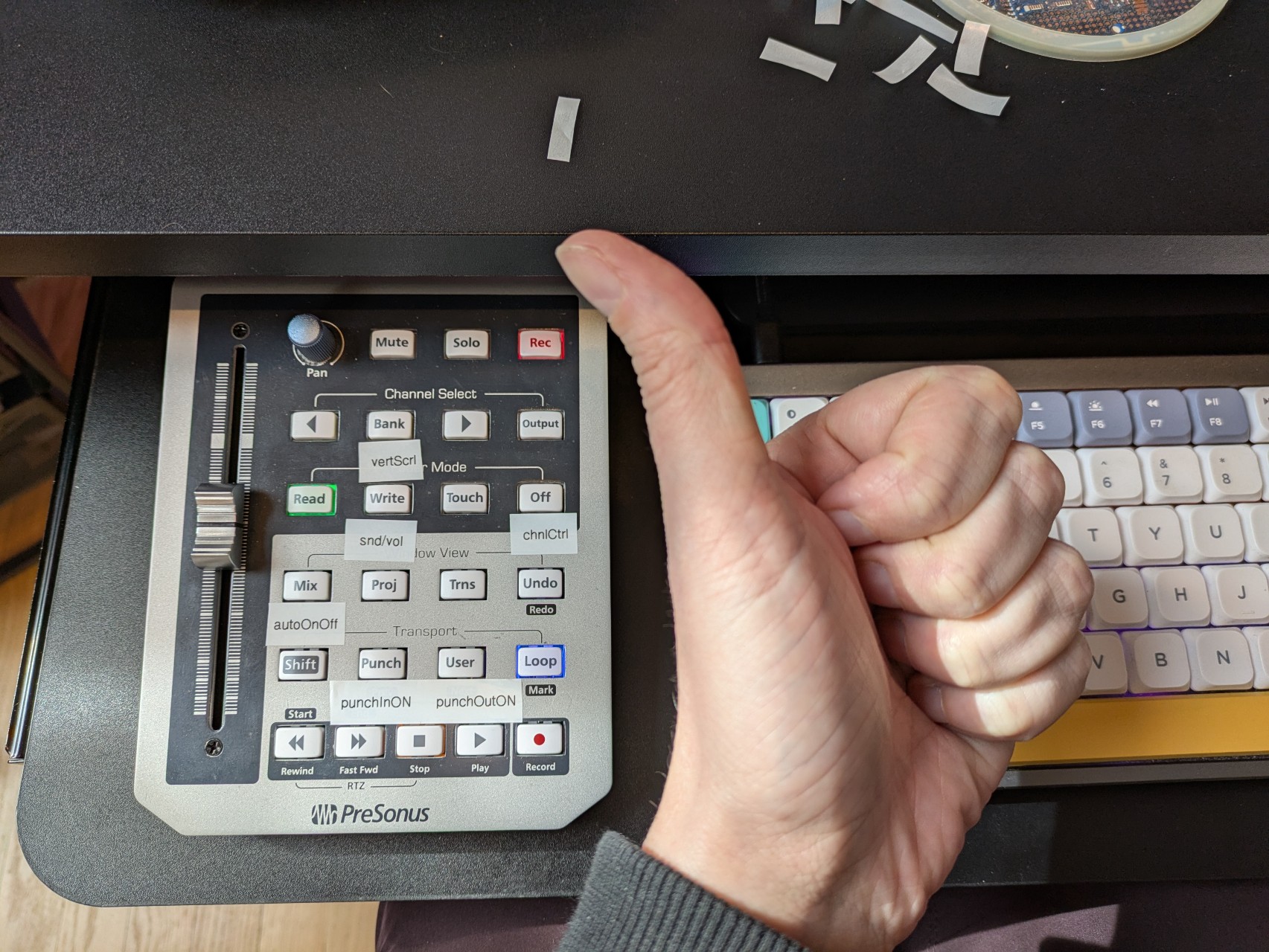 thumbs up in front of a Faderport classic next to a mechanical keyboard. The Faderport has extra labels for Bitwig functions.